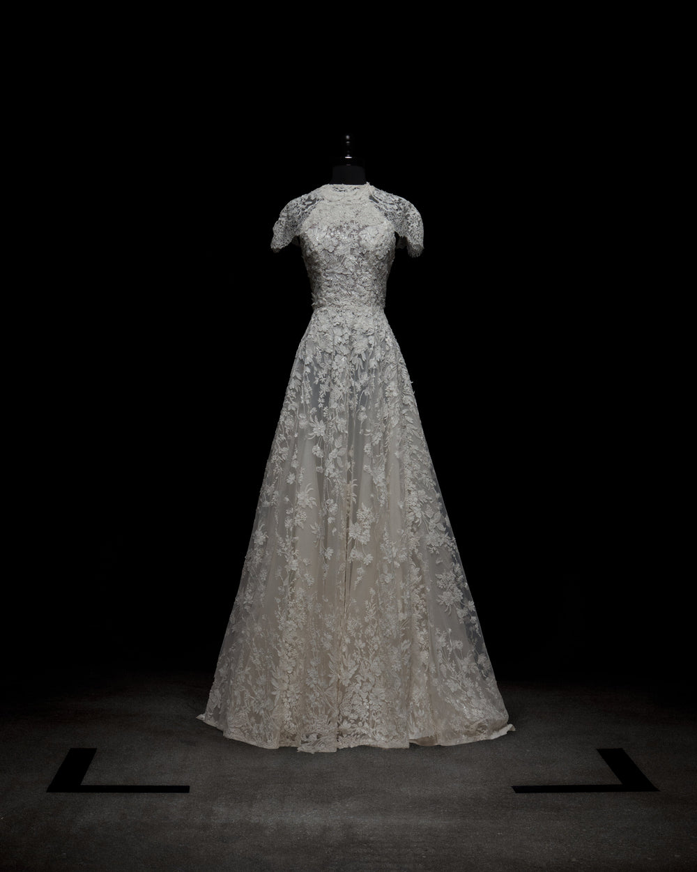 The Leia Dress with Cape by SVT DESIGNS, a white lace floor-length gown with short sleeves and a fitted bodice, is showcased on a black mannequin against a dark background. Black markers form a square on the floor around the dress.