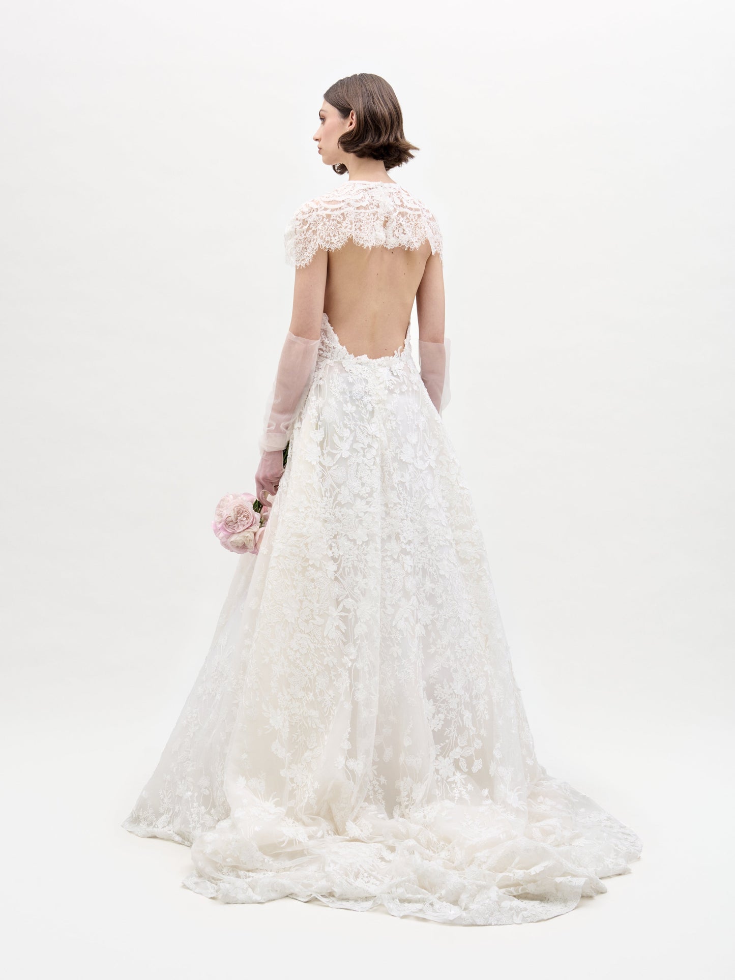 A woman with short brown hair, wearing the SVT DESIGNS Leia Dress with Cape featuring a low back and sheer long sleeves, stands facing away holding pink flowers against a plain white background.