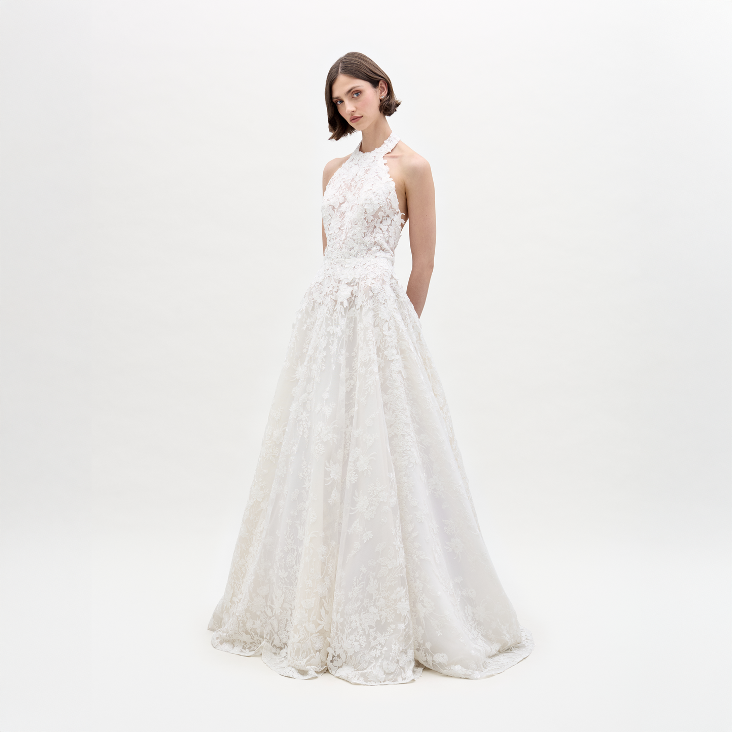 A woman with short brown hair stands against a white background wearing the Leia Dress by SVT DESIGNS—a sleeveless, floor-length white lace maxi with a fitted bodice and full skirt, crafted from fine Italian fabric. Her hands are held behind her back.