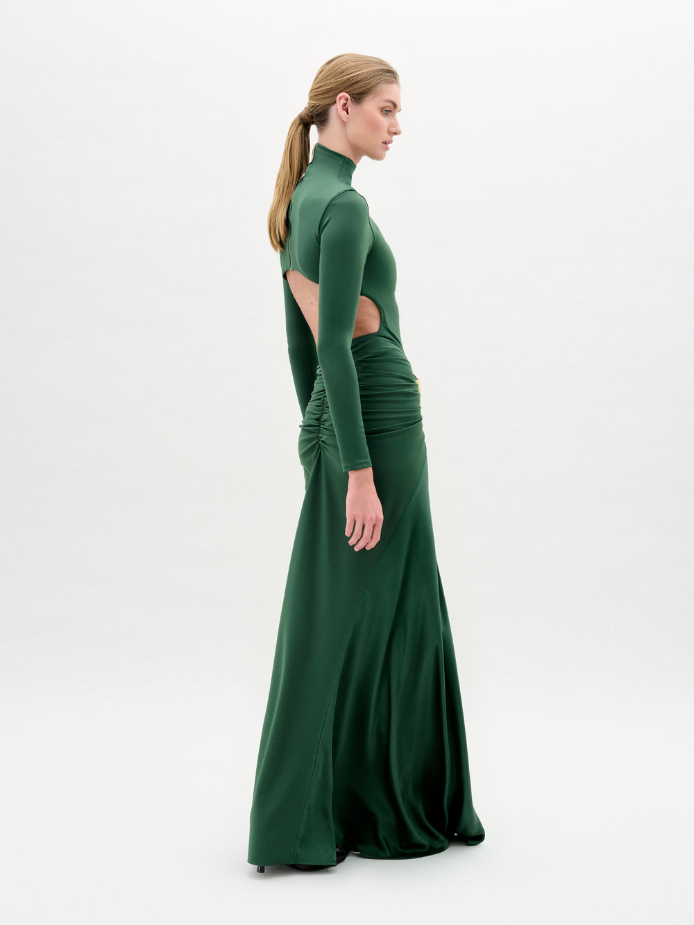 A person with long hair tied back wears the SVT DESIGNS Lena Dress, an emerald green maxi with a high neck and long sleeves. Gold embellishments and side cut-outs adorn it, while its ruched design stands out against a plain white backdrop.