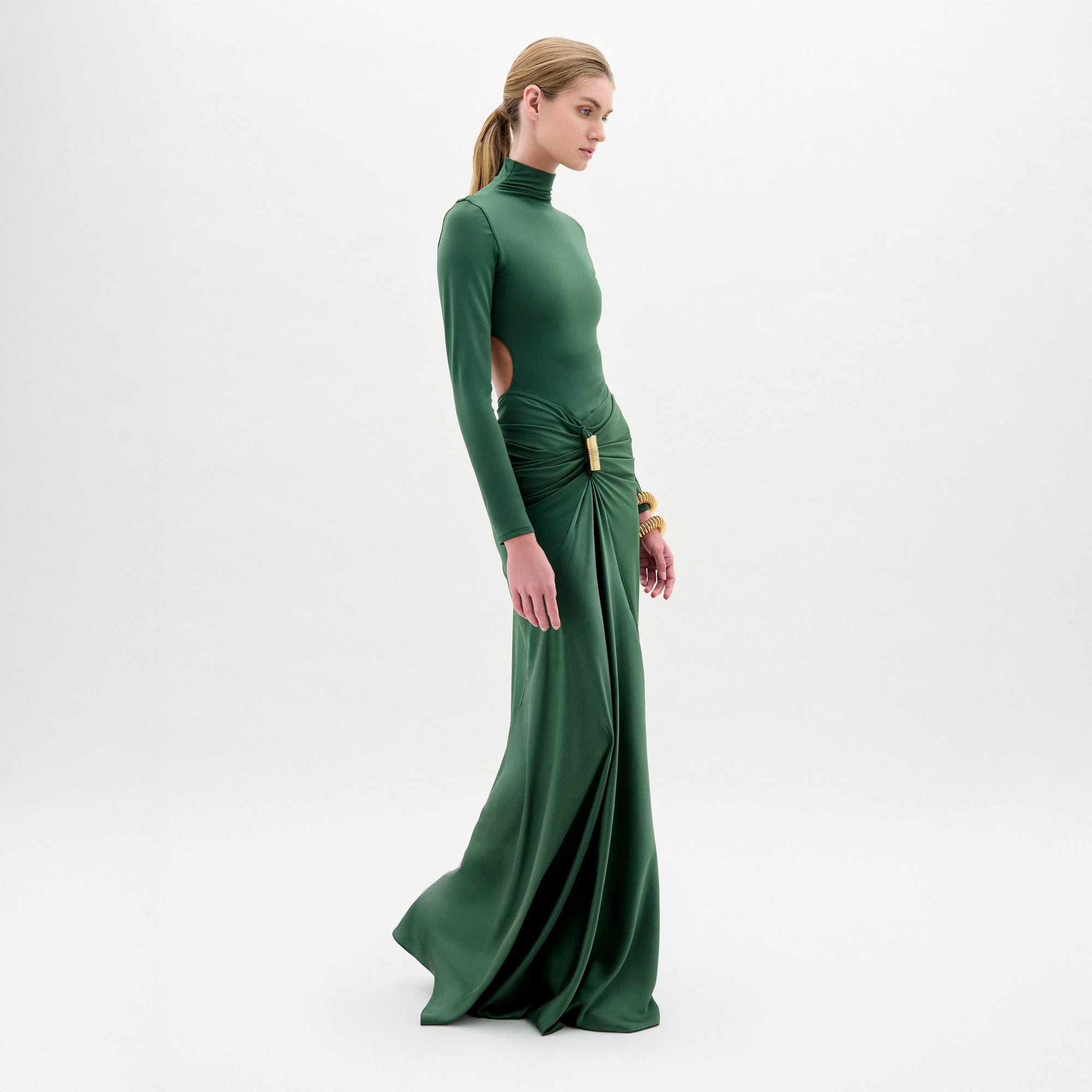 A woman wears the Lena Dress—a long green maxi with a high neck, gathered skirt, long sleeves, and side cutouts—while standing against a plain white background.