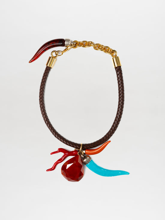 The SVT DESIGNS Leslie Choker features a braided leather design with a gold chain clasp, adorned with multicolor charms including a red chili pepper, blue horn, and red bead with branch-like protrusions, all highlighted by metallic accents.