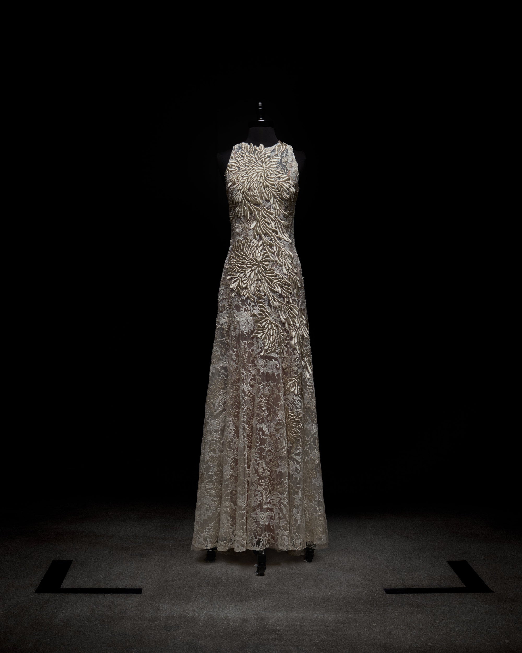 On a mannequin against a dark backdrop, the Levi Dress by SVT DESIGNS is full-length and sleeveless. It features intricate floral lace detailing throughout, a high neckline, and is light-colored.