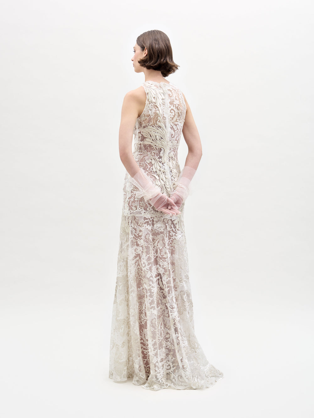 A woman with short hair faces away, showcasing the Levi Dress by SVT DESIGNS—a long, sleeveless lace dress featuring intricate patterns. Her hands are lightly clasped behind her back against a plain white background.