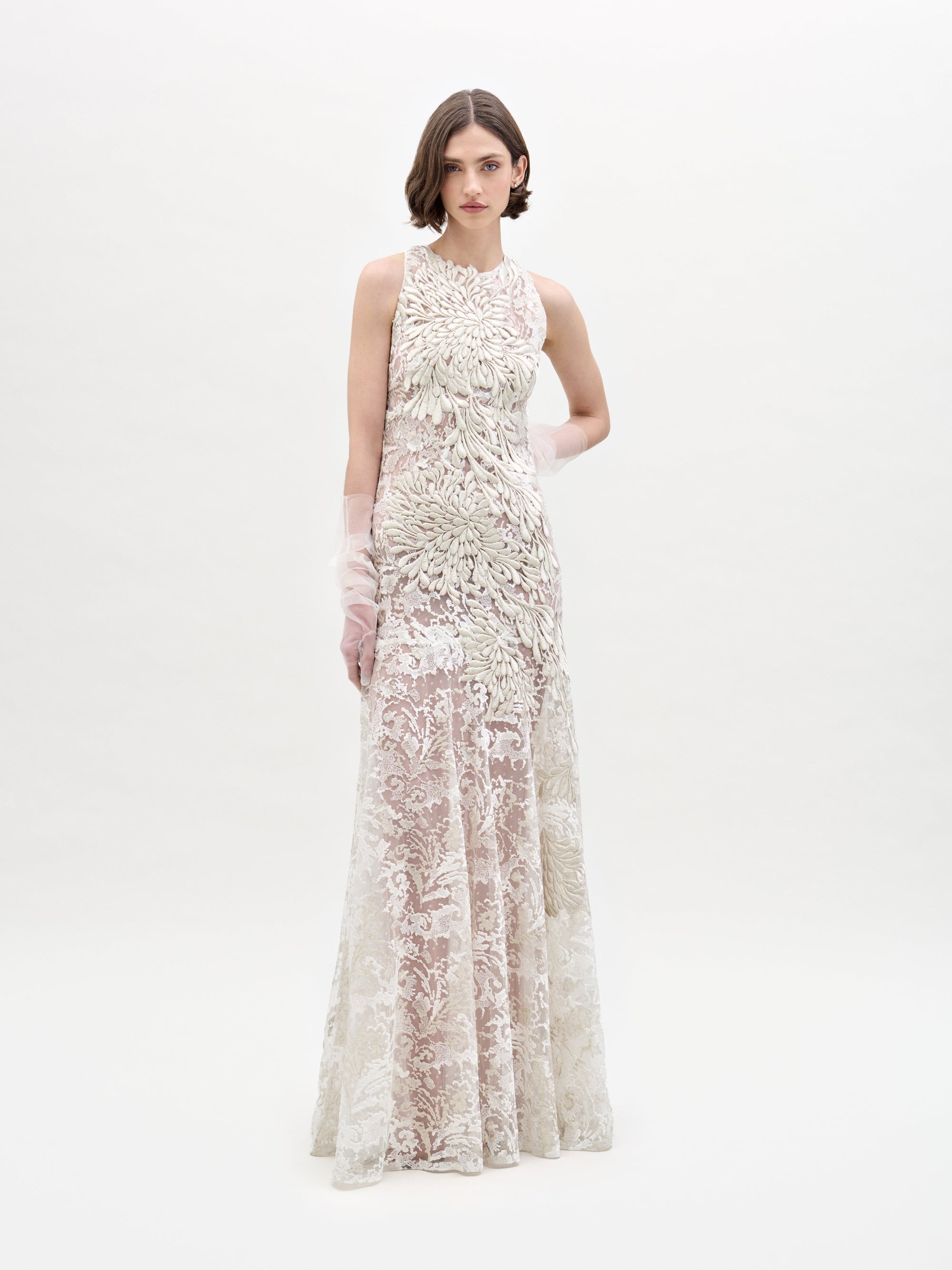 Against a plain background, a person wears the Levi Dress by SVT DESIGNS, a long, sleeveless lace gown with intricate floral patterns. Their short wavy hair complements the pose of an arm across their waist.