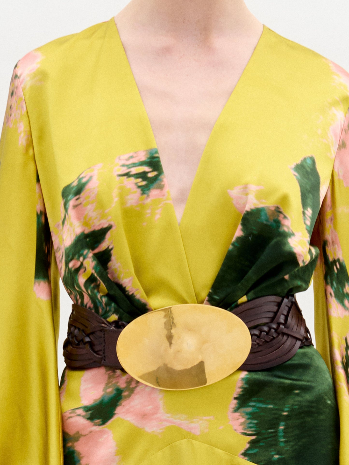 A person wears the Lima Dress by SVT DESIGNS—a yellow maxi with green and pink abstract print, a deep V-neckline, and a wide dark belt featuring a large oval gold buckle at the waist.
