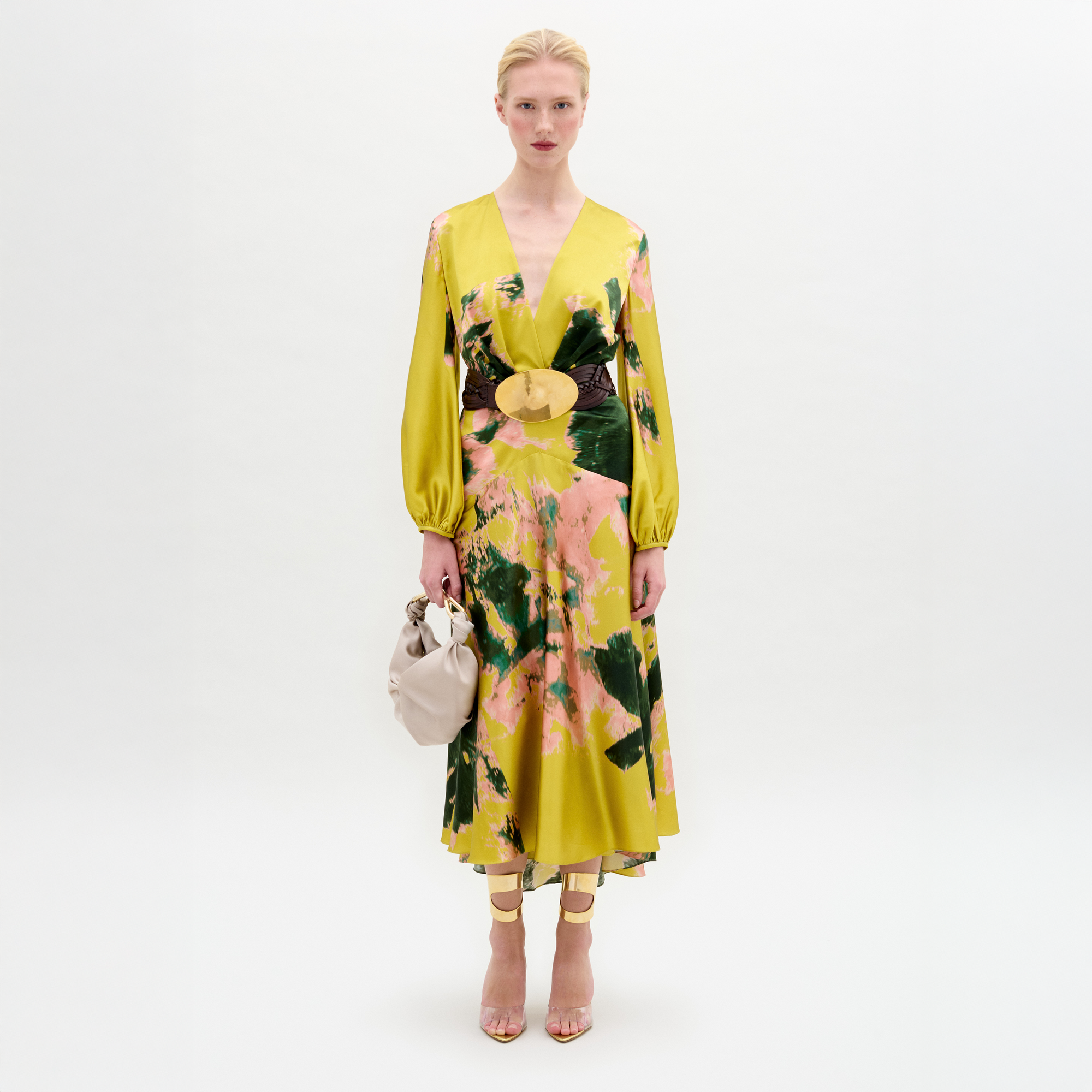A person stands against a white background wearing the Lima Dress by SVT DESIGNS, a yellow and green floral maxi with long sleeves, styled with a wide brown belt, nude heels, and a beige handbag.
