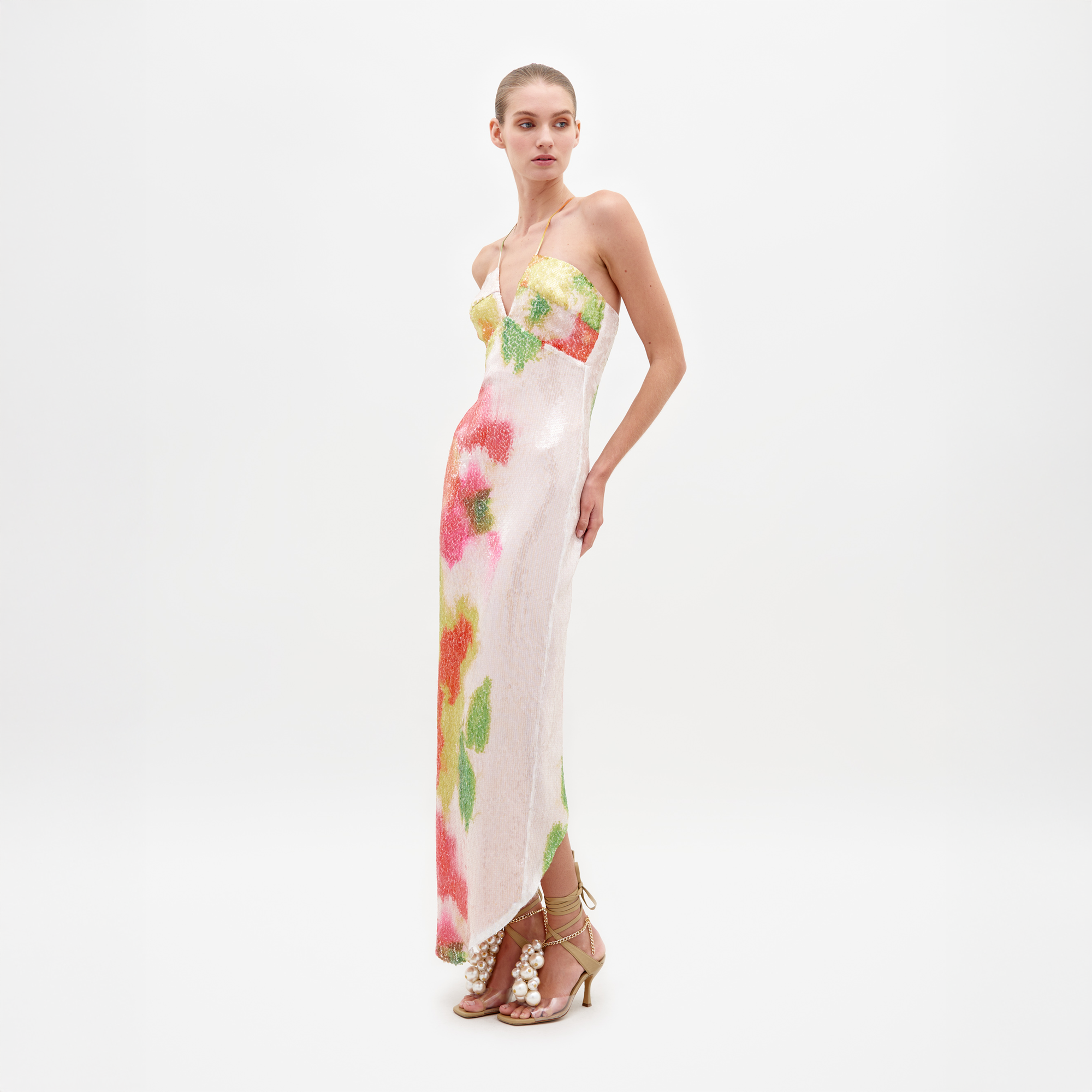 A woman is wearing the Lisseth Dress Spring Blooms, a strapless white dress adorned with colorful floral patterns, paired with high-heeled sandals featuring ankle straps and large pearl embellishments—an ideal choice for those who pre-order by February 15th to ensure timely shipping.