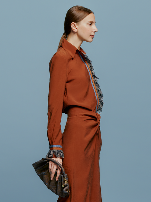 A person stands against a blue background wearing the Liv Blouse Cacao, a rust-colored outfit with fringe details on the collar, front, cuffs, and waistband, highlighting the latest trend expected in the September 2nd shipment. They're holding a small gray handbag.