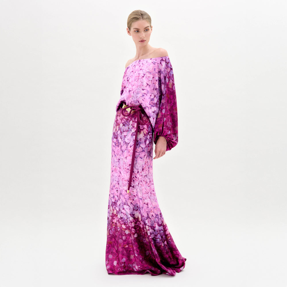 A woman stands against a white background wearing the Lorella Dress by SVT DESIGNS—an off-the-shoulder maxi in luxurious Italian fabric, featuring a pink and purple abstract pattern and a matching waist belt. She gazes slightly left.