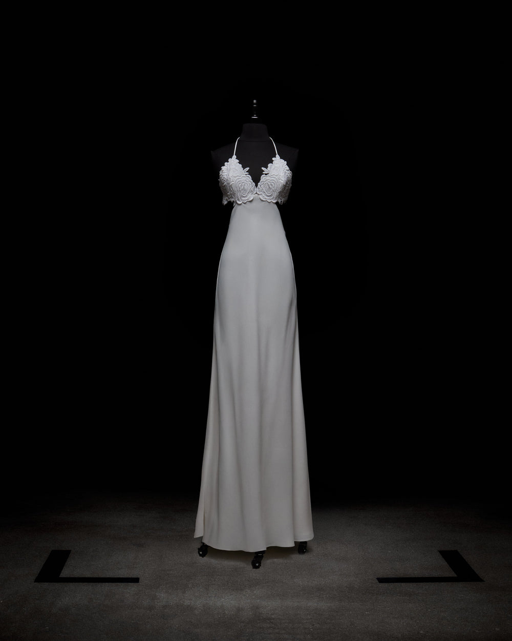 The Lua Dress by SVT DESIGNS, showcased on a mannequin against a black background, is an elegant white long dress with a fitted bodice, intricate floral detailing, and a halter-style neckline that flows into a smooth skirt.