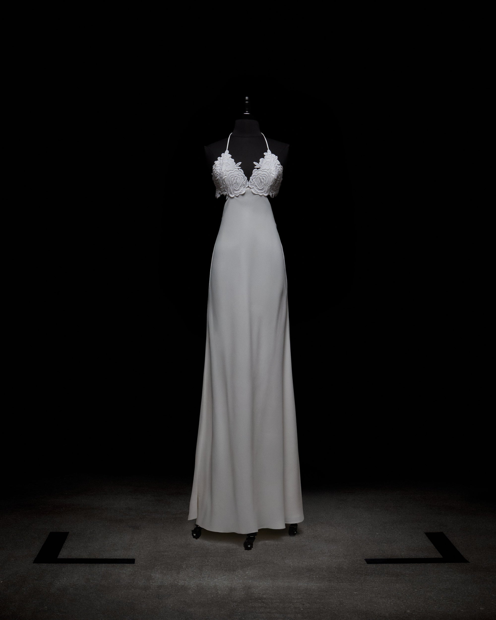 The Lua Dress by SVT DESIGNS, showcased on a mannequin against a black background, is an elegant white long dress with a fitted bodice, intricate floral detailing, and a halter-style neckline that flows into a smooth skirt.