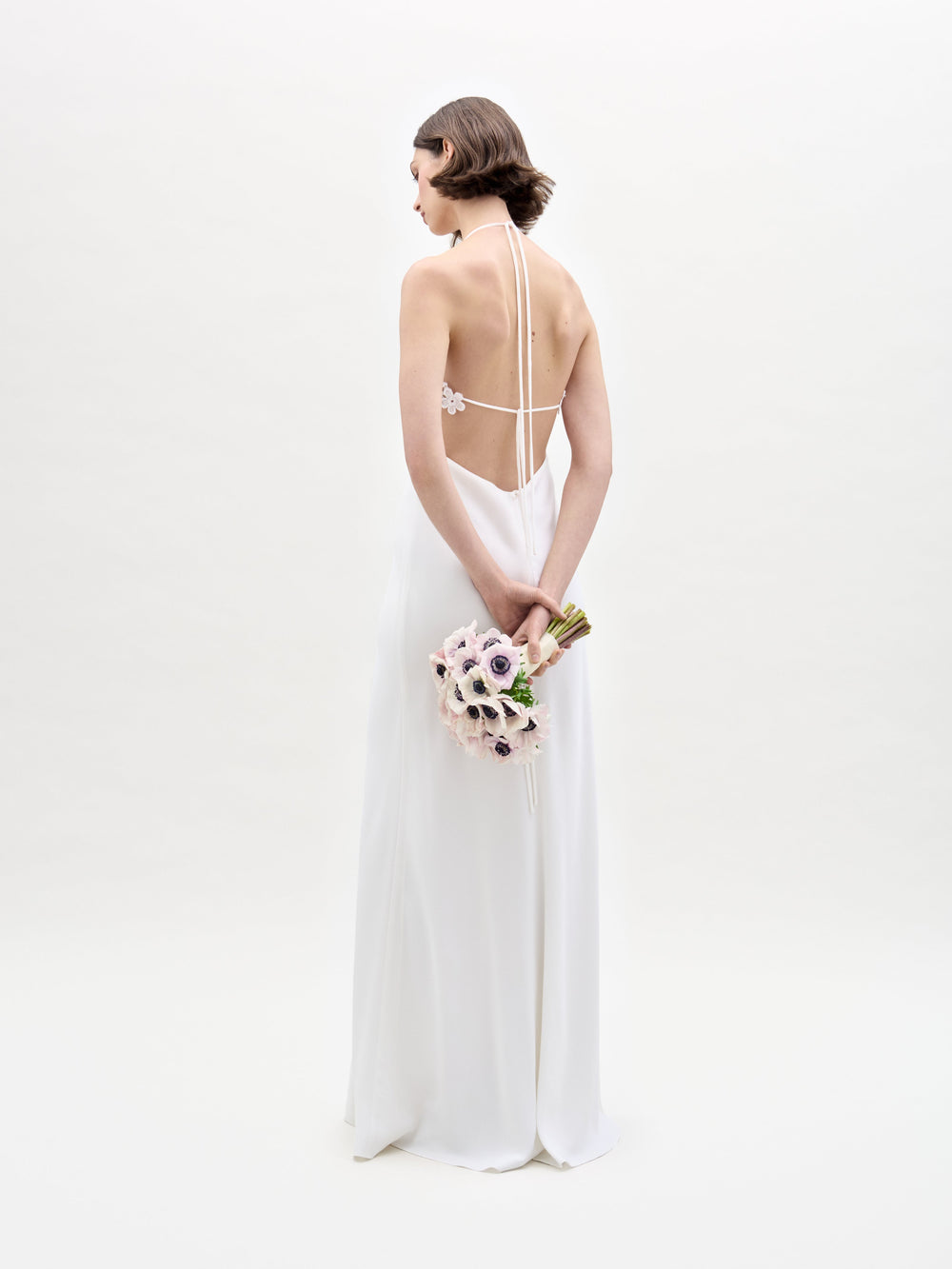 In a plain white backdrop, a woman showcases her back to the camera in SVT DESIGNS' Lua Dress, a backless white gown. She holds a bouquet of light-colored flowers with dark centers.