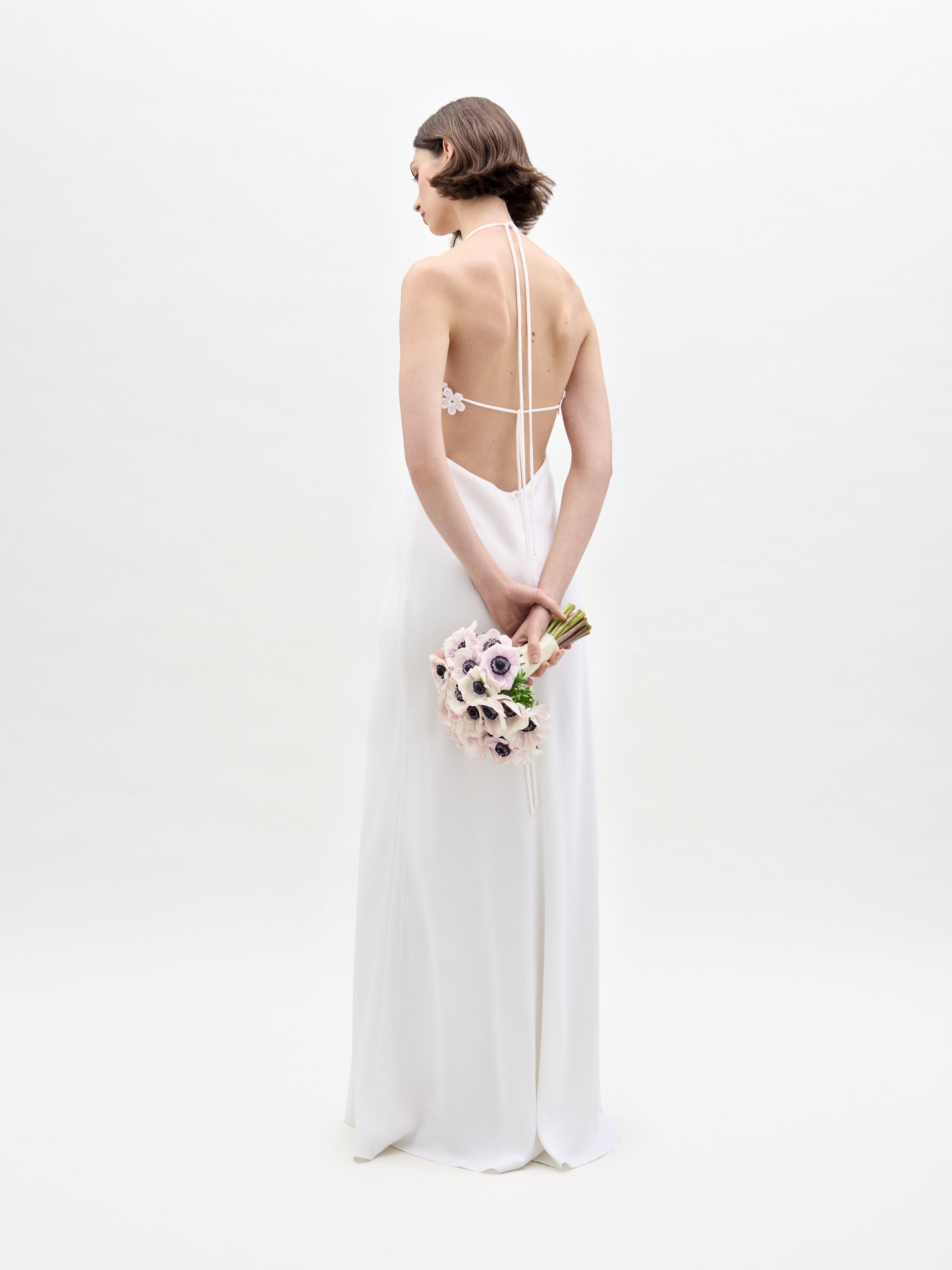 In a plain white backdrop, a woman showcases her back to the camera in SVT DESIGNS' Lua Dress, a backless white gown. She holds a bouquet of light-colored flowers with dark centers.