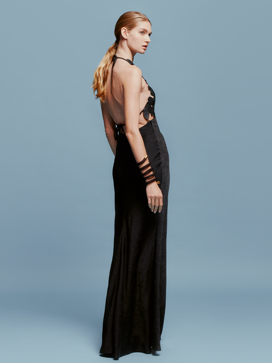 A person stands against a blue background, wearing the elegant Lua Dress Black, adorned with large black flowers on the bodice, capturing the timeless essence of high fashion.