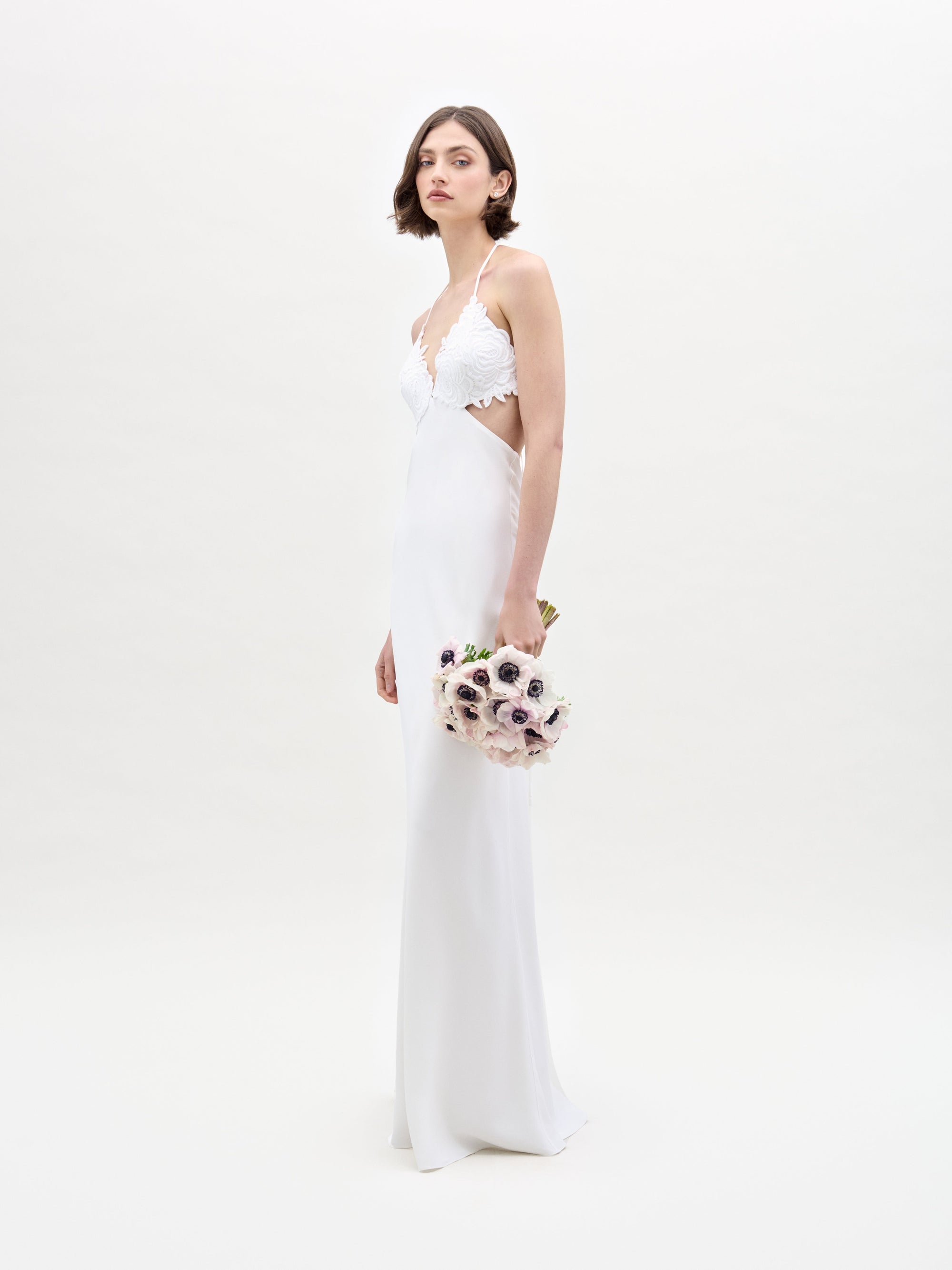 A woman poses against a white background in the SVT DESIGNS Lua Dress, featuring lace details. She holds a bouquet of flowers at her side, with her dark hair styled in a bob.