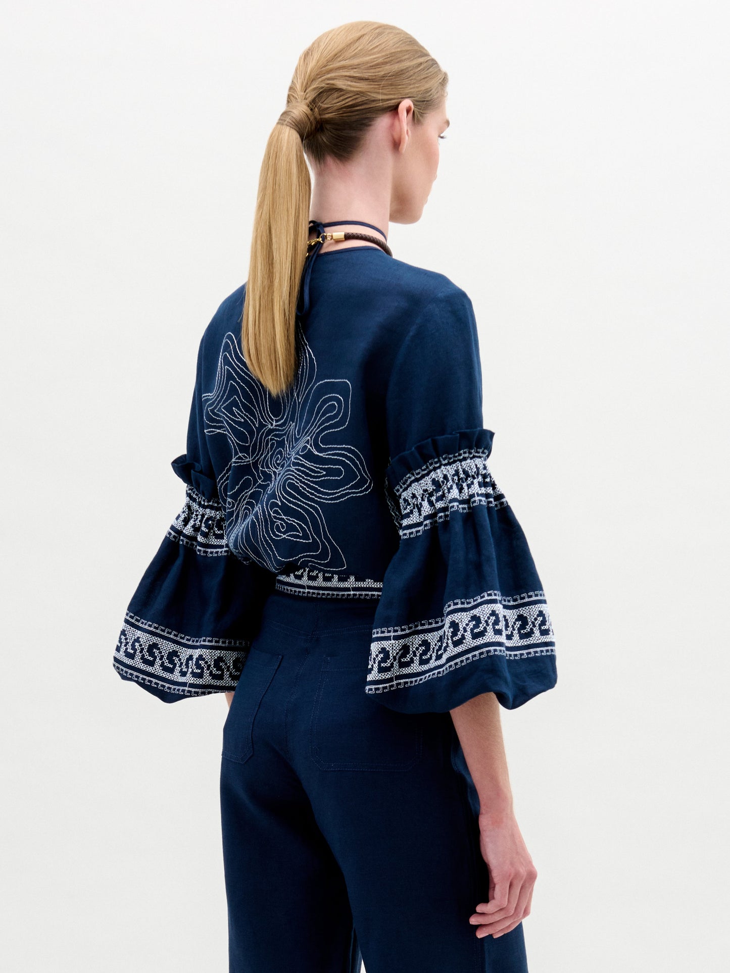 A person with long blonde hair in a ponytail wears the SVT DESIGNS Lucaya Blouse, a navy blue Italian fabric piece. It has voluminous sleeves with detailed white embroidery and a large embroidered design on the back against a plain white background.