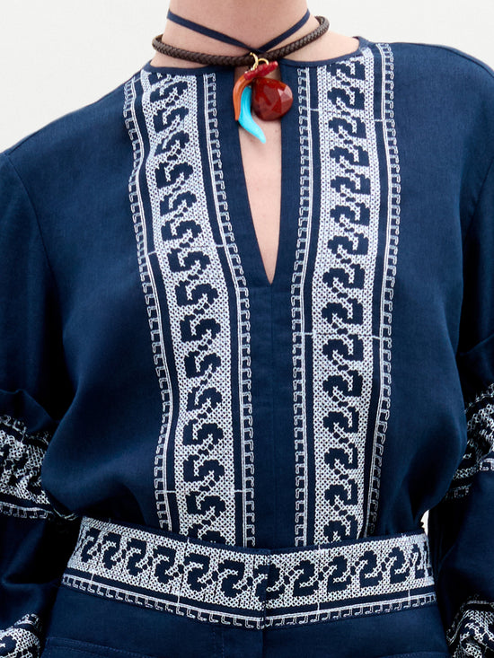 A person wearing SVT DESIGNS' Lucaya Blouse, a dark blue traditional garment made from luxurious Italian fabric with intricate white embroidery and voluminous sleeves. A necklace with a dark cord and glossy red pendant elegantly adorns their neck.