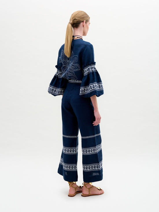 Dressed in SVT DESIGNS' Zina Pant, a person with long hair tied back stands in a navy blue ensemble with intricate white patterns, crafted from fine Italian fabric. The outfit features wide, ruffled sleeves and a wide-leg silhouette. They wear sandals while facing away from the camera against a plain background.