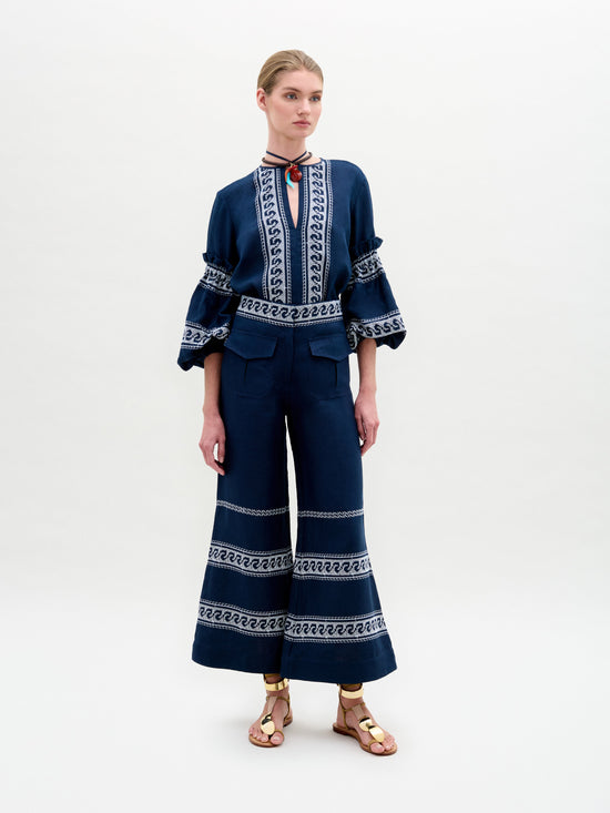 A person is posing against a plain backdrop, wearing a blue outfit featuring white geometric patterns. The top has ruffled sleeves, and the wide-leg silhouette pants are SVT DESIGNS' Zina Pant made from Italian fabric. Brown sandals complete the look, with hair pulled back and hands relaxed.