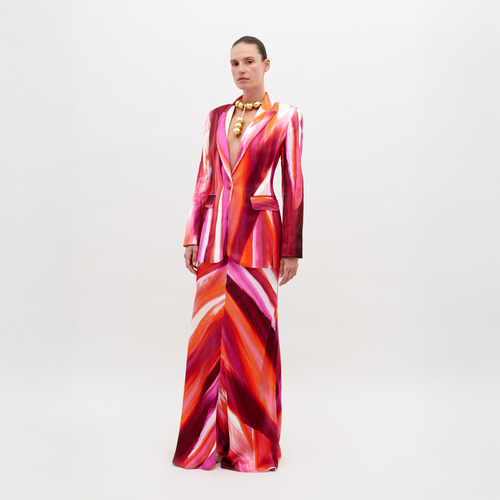 A person in a Laurina Skirt by SVT DESIGNS stands before a plain white background, wearing a boldly patterned Italian textile suit in red, pink, orange, and white. They sport short hair and a large statement necklace.