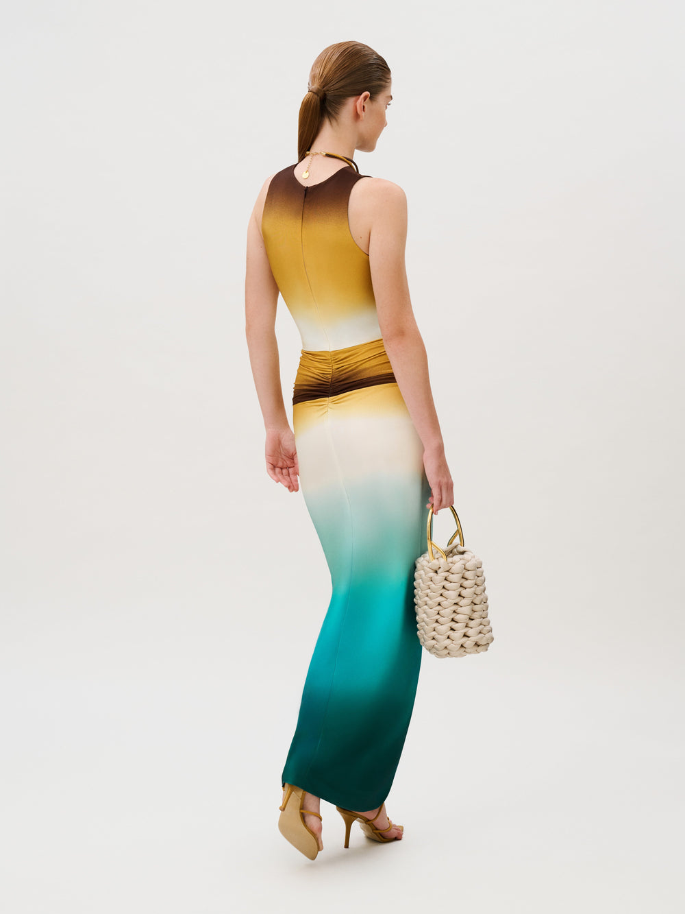 A person wearing the Leni Dress, a slim-fit, floor-length gown with a horizontal ombré gradient from brown to yellow, cream, and deep teal, carrying a cream-colored woven handbag.