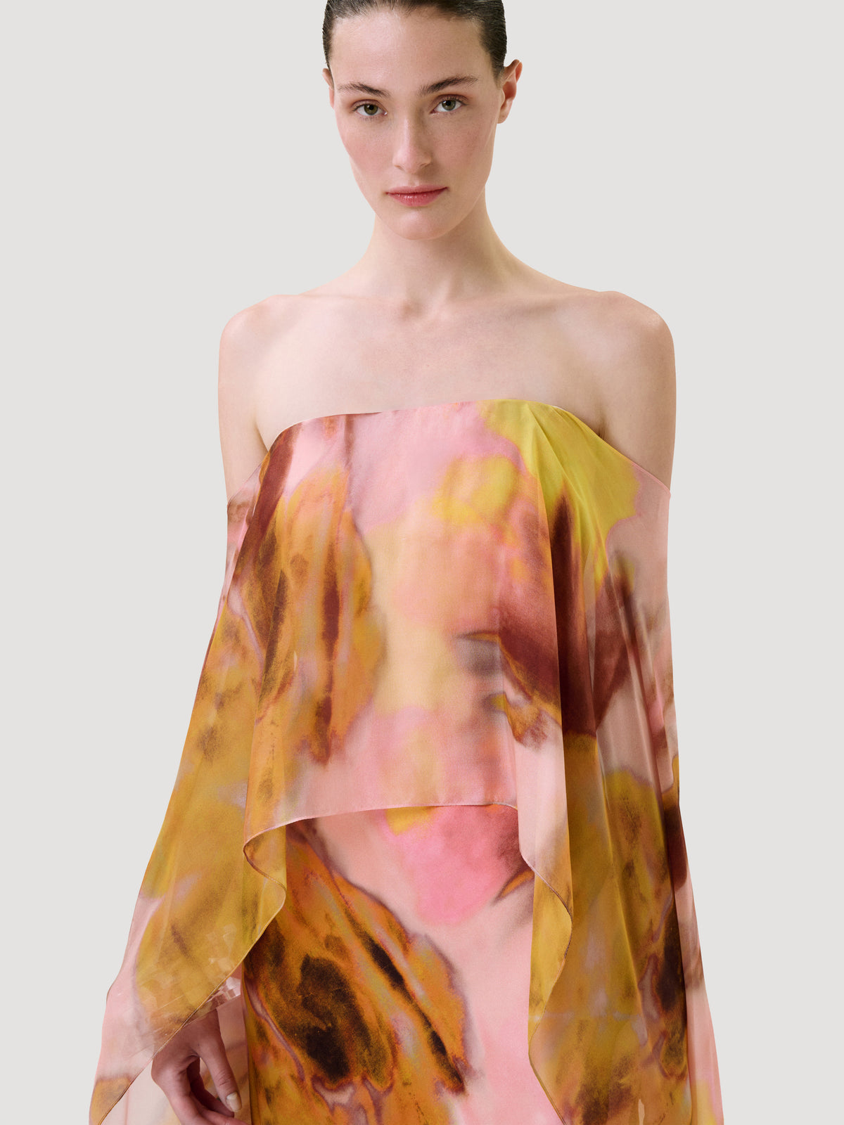 A model wearing a sheer, strapless draped wrap or dress in a pink, yellow, and orange abstract floral print against a light gray studio background.