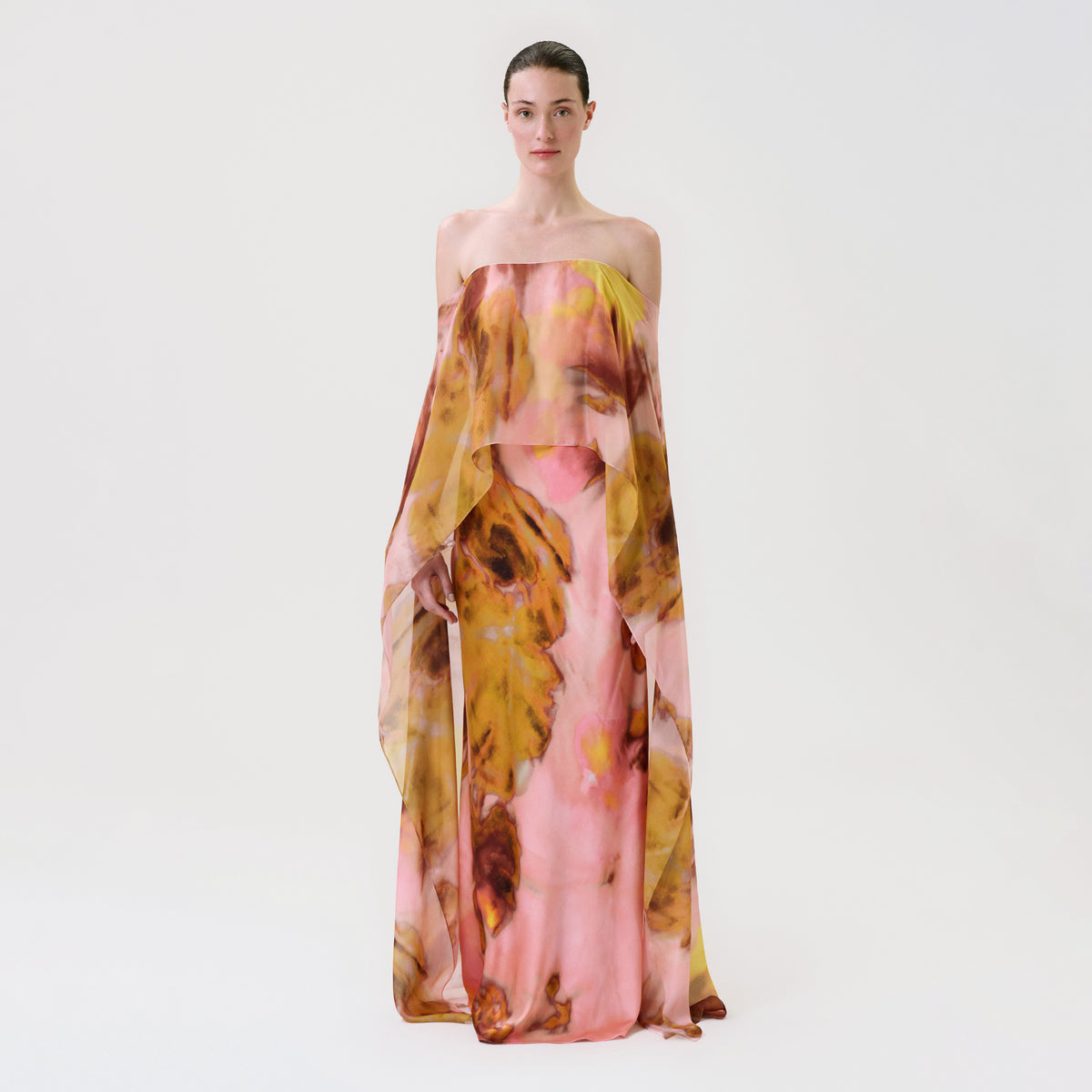 A model stands against a plain white studio background wearing a strapless, floor-length sheer gown with an abstract floral print in pink, mustard yellow, and brown tones.