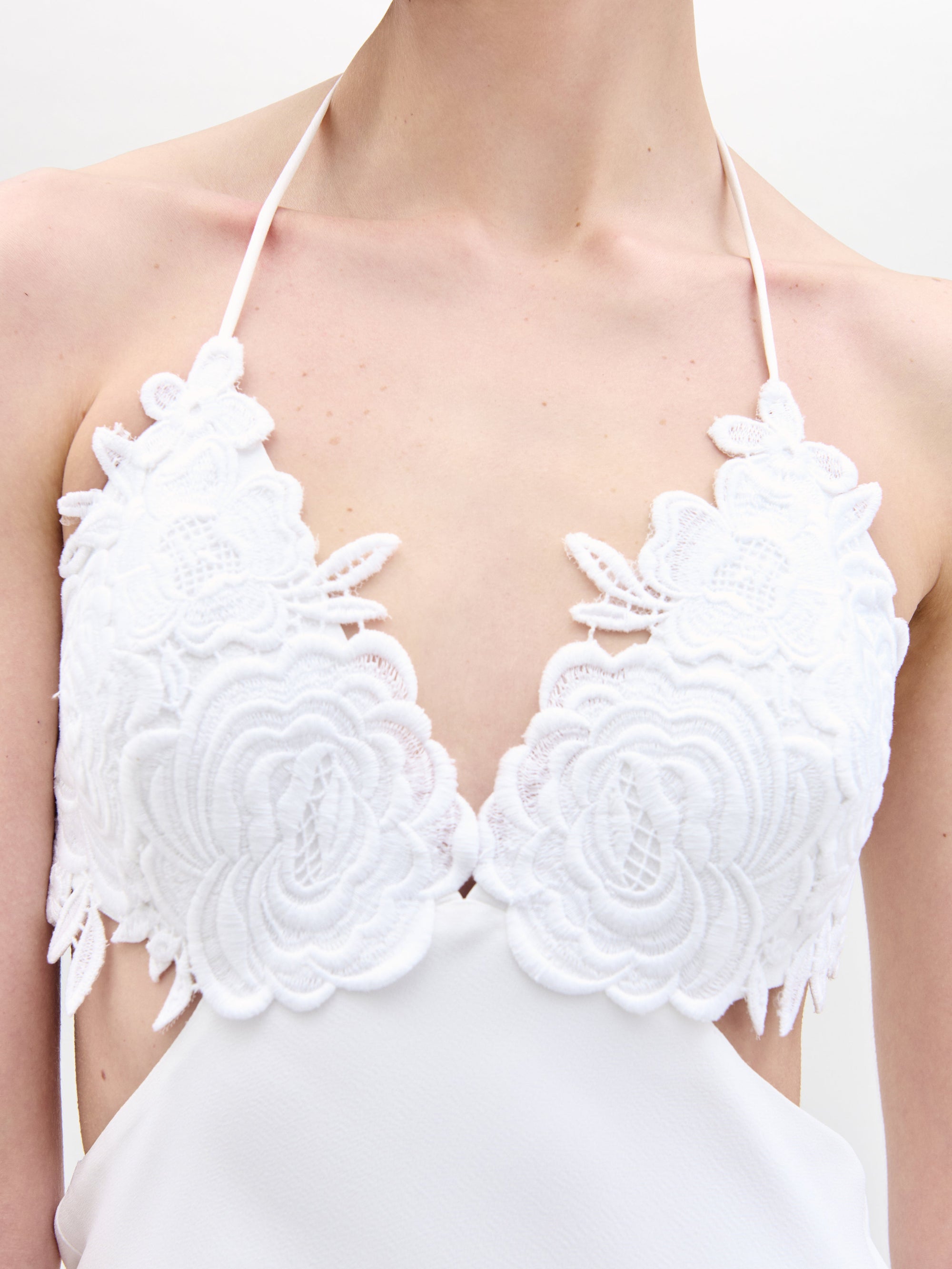 Close-up of someone in a Lua Dress by SVT DESIGNS, featuring a white halter-top with floral lace around the neckline. The background is plain and blurred.