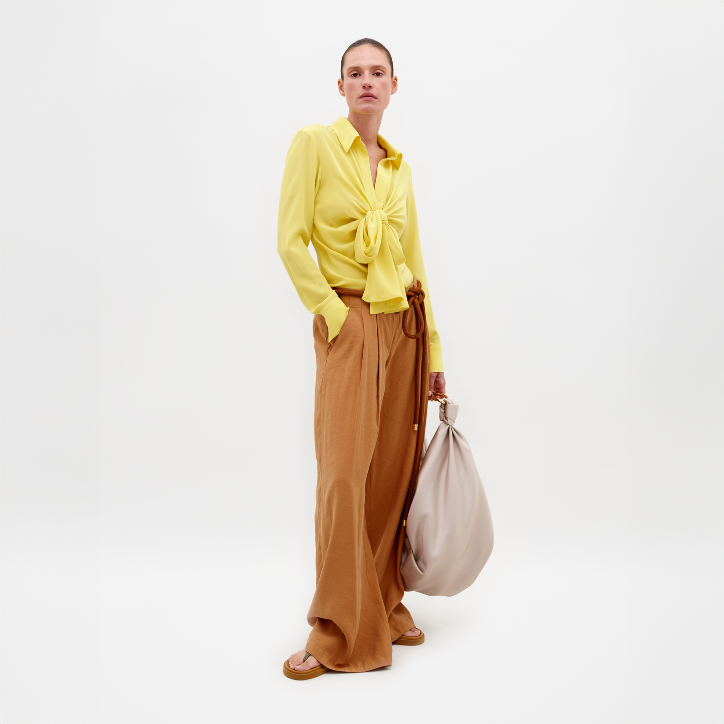 Against a white backdrop, a person wears SVT DESIGNS' yellow tie-front blouse paired with the Opal Pant, a high-waisted, wide-leg design. A large beige bag is in one hand while the other rests in their pocket. Made in Colombia, brown sandals complete the look.