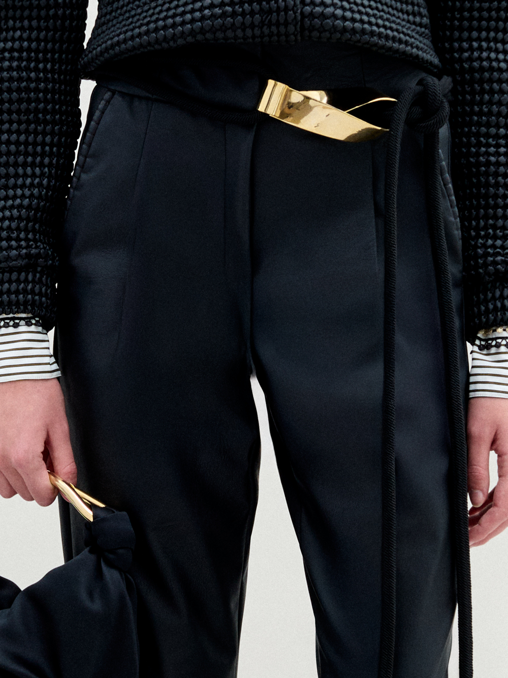 A person wears the SVT DESIGNS Moad Pant—black, high-waisted, cropped with a straight leg in Italian fabric—styled with a textured black top and gold-accented accessories against a plain, light background.