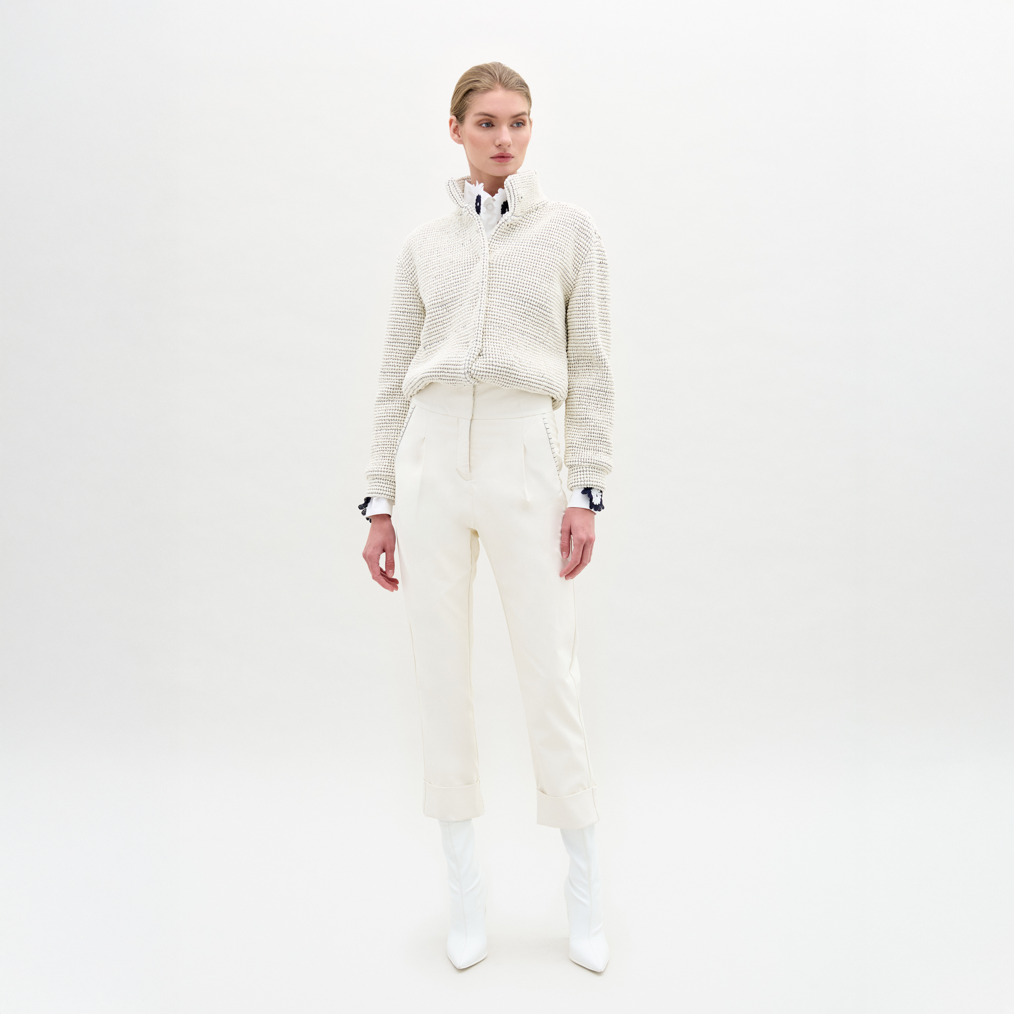 A person faces forward with hands at their sides, wearing a textured white sweater over a collared shirt, SVT DESIGNS Moad Pant—high-waisted and cropped in Italian fabric—and white pointed boots against a plain white background.
