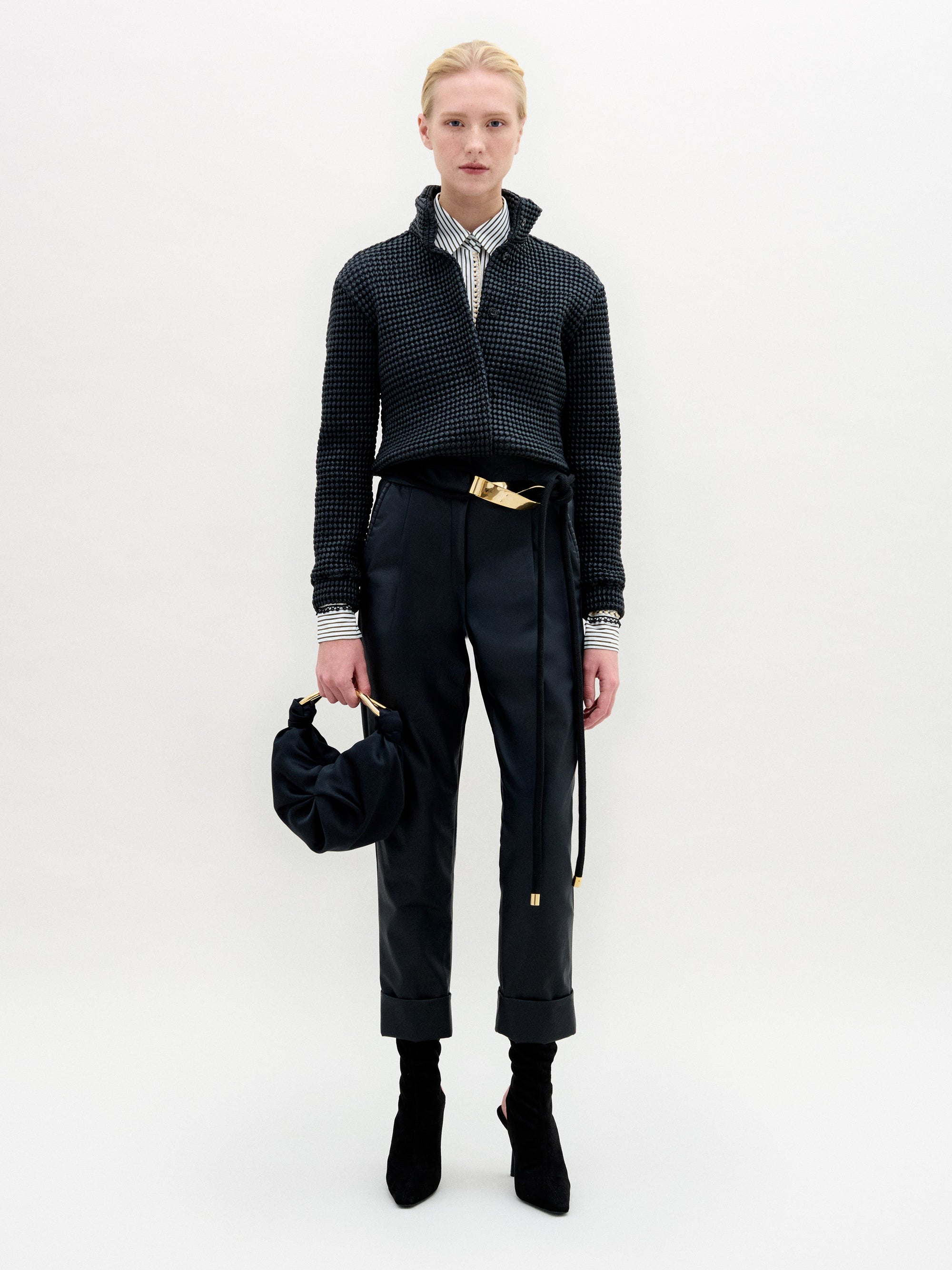 A person with blond hair stands against a white background, wearing SVT DESIGNS' Moad Pant—a high-waisted cropped style with a belt—paired with a dark jacket, striped shirt, heeled boots, and holding a dark handbag.