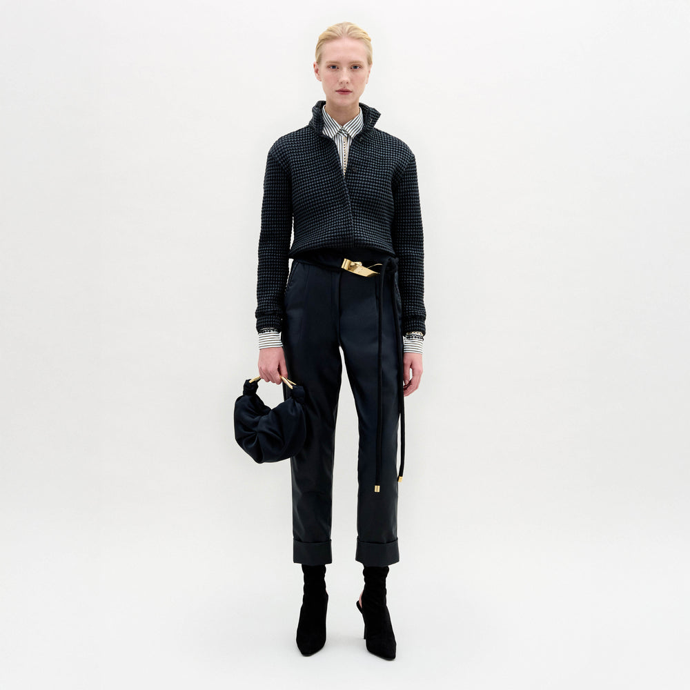 A person stands against a plain white background wearing SVT DESIGNS’ Moad Pant—straight fitted black trousers crafted from fine Italian fabric—paired with a dark textured jacket, collared shirt, black boots, and holding a small black handbag.