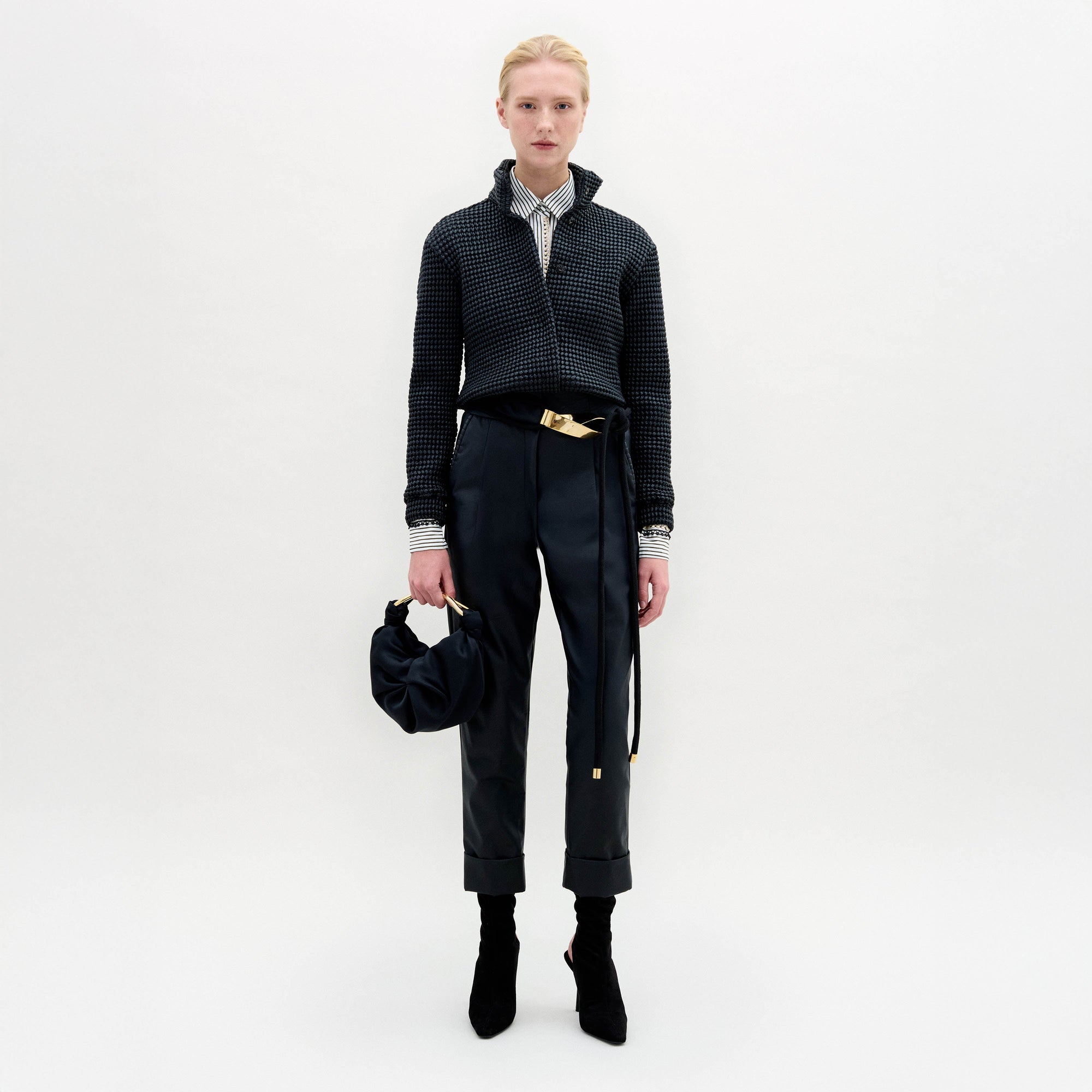 Person stands against a plain white background wearing the Moad Pant, a dark textured jacket, patterned shirt, black boots, and holding a small black handbag.