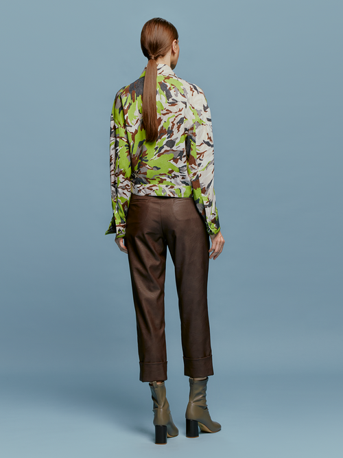 A person is standing against a blue background, wearing a green and brown patterned shirt, dark pants referred to as the "Moad Pant Brown," and gray heeled boots. The shirt is partially tucked into the pants, creating a stylish look perfect for August 19th.