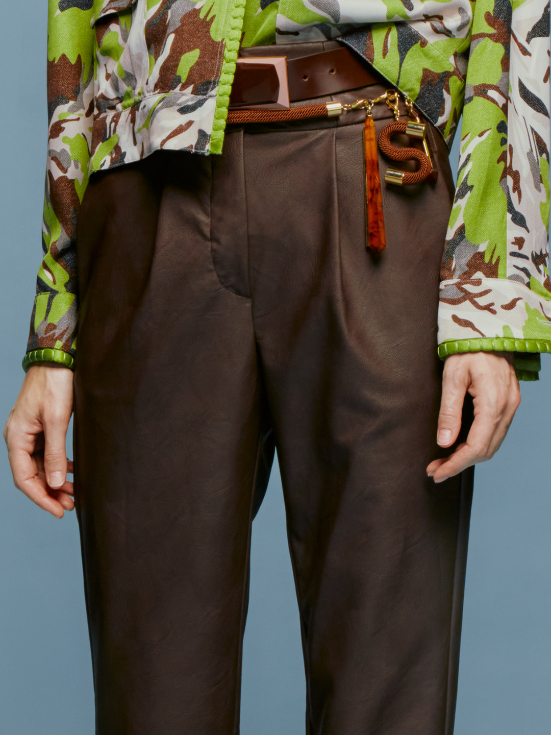 A person is standing against a blue background, wearing a green and brown patterned shirt, dark pants referred to as the "Moad Pant Brown," and gray heeled boots. The shirt is partially tucked into the pants, creating a stylish look perfect for August 19th.