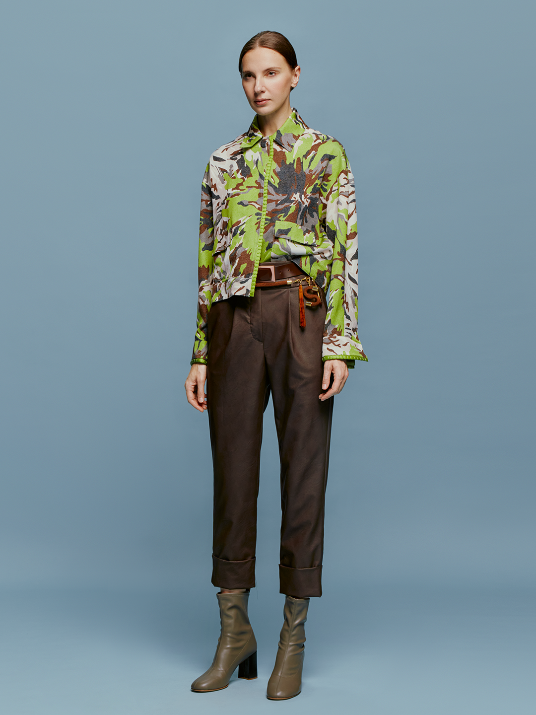 A person is standing against a blue background, wearing a green and brown patterned shirt, dark pants referred to as the "Moad Pant Brown," and gray heeled boots. The shirt is partially tucked into the pants, creating a stylish look perfect for August 19th.