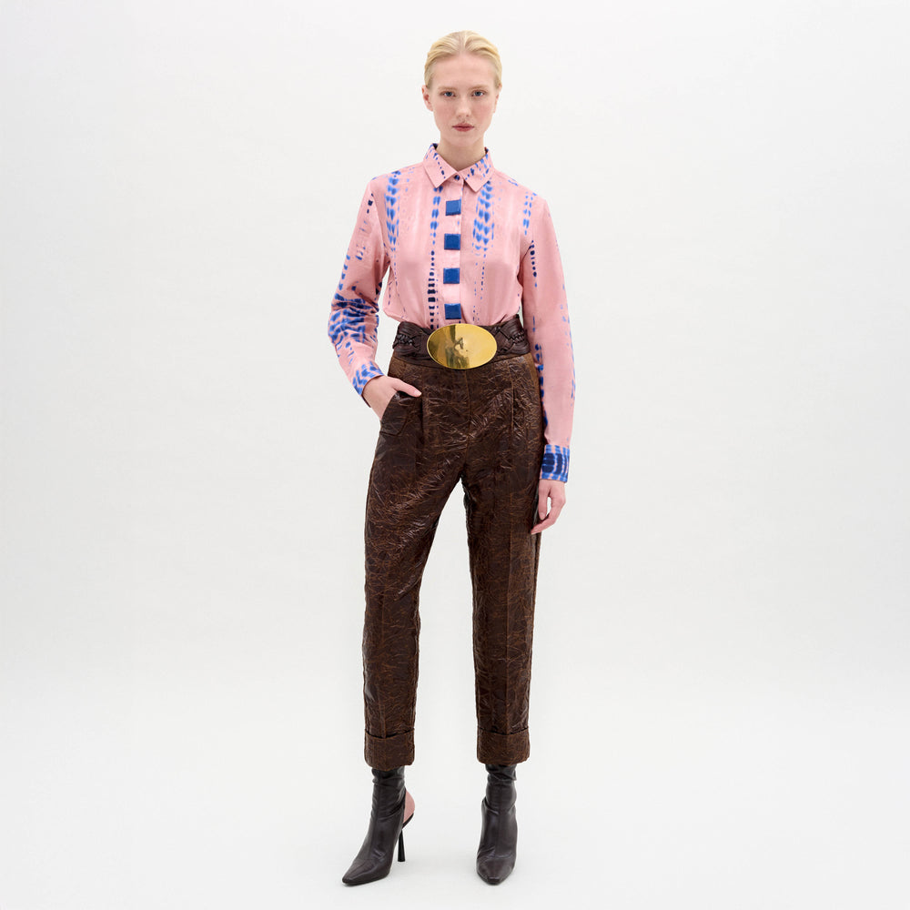 A person stands against a plain white background wearing SVT DESIGNS’ Moad Pant—high waisted, straight fitted, cropped Italian fabric trousers—with a pink patterned blouse, wide gold belt, and dark heeled boots. One hand is in their pocket.