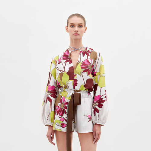 A person stands against a plain white background, wearing the Molveno Blouse by SVT DESIGNS. The floral V-neck blouse features pink, green, and brown flowers, paired with white shorts and a brown belt. Their hair is pulled back as they look directly at the camera.