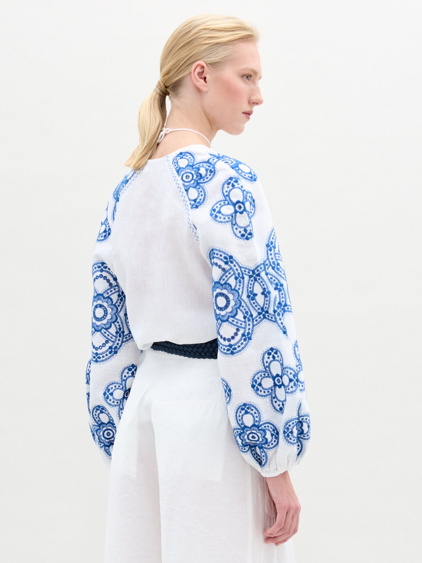 A person with light hair faces sideways, styled in the Molveno Blouse by SVT DESIGNS. The blouse showcases blue floral embroidery on the sleeves and back, paired elegantly with a black belt against a plain white background.