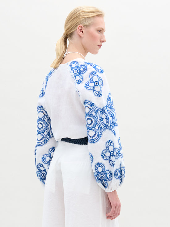 A person with light hair faces sideways, styled in the Molveno Blouse by SVT DESIGNS. The blouse showcases blue floral embroidery on the sleeves and back, paired elegantly with a black belt against a plain white background.