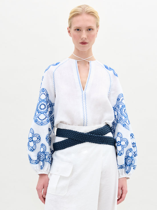 A person with blond hair wears the SVT DESIGNS Molveno Blouse featuring blue floral embroidery on the sleeves and a V-neckline against a white background. They pair it with Italian-fabric white pants and cinch their waist with a dark woven belt.