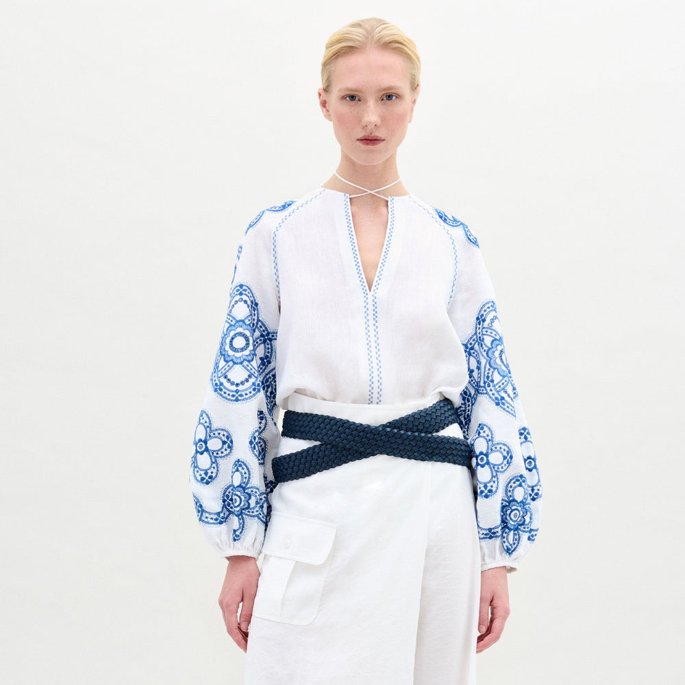 A woman with blonde hair in a low bun wears the Molveno Blouse—an embroidered linen top with gathered sleeves and a deep V neckline—paired with a thick navy braided belt and a white cargo-pocket skirt, against a plain background.