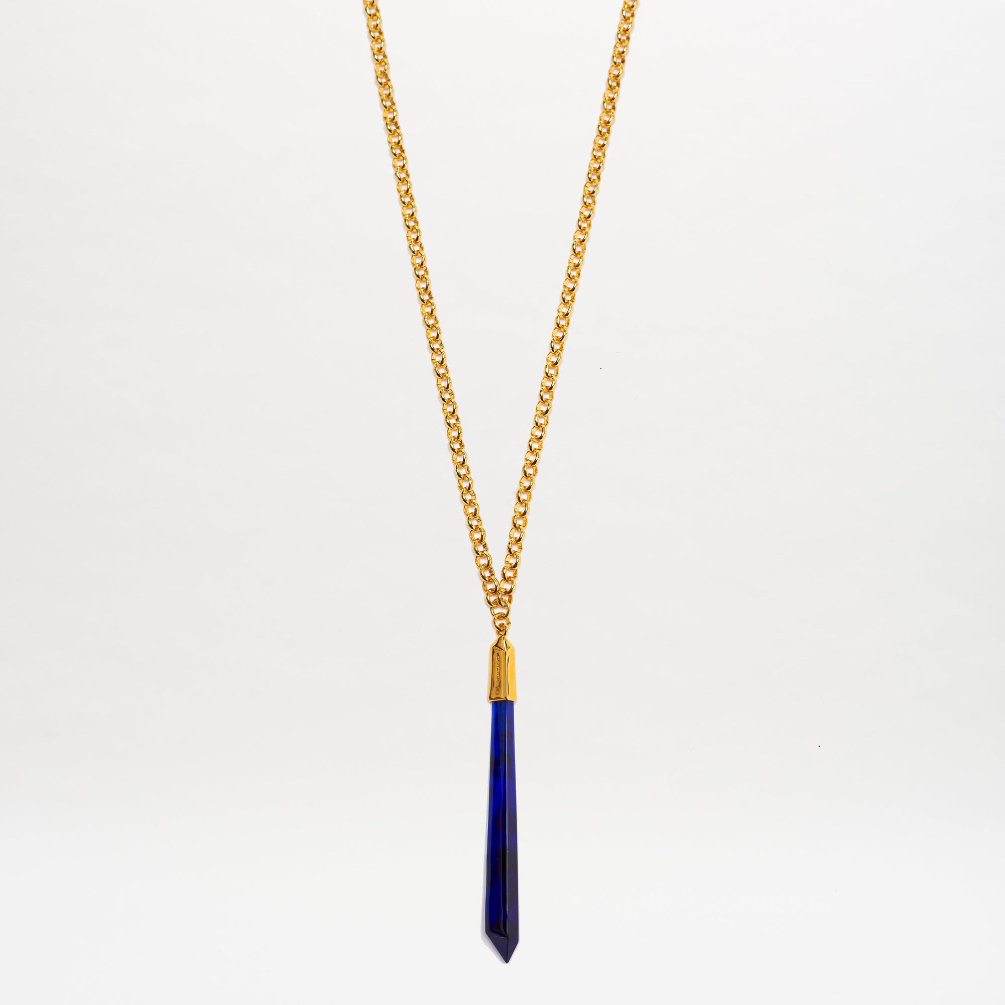 The Montse Necklace by SVT DESIGNS features a gold chain with a long, narrow dark blue geometric crystal pendant, shown hanging vertically against a plain white background.