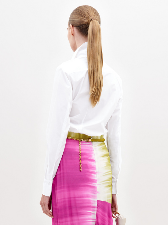 A person wearing a Mercury Blouse White and a vibrant pink skirt with green accents, standing against a plain white background, available for pre-order now. Ships by November 15th, 2024.