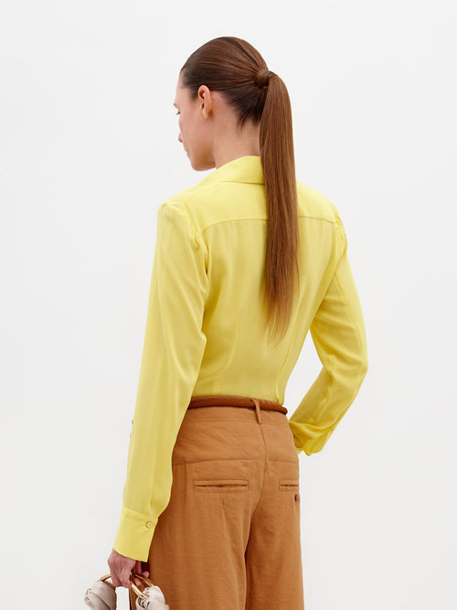 An individual stands against a plain backdrop wearing the Mercury Blouse Yellow, featuring long sleeves and a knotted front, paired with light brown belted trousers. They hold a small beige bag in their left hand. Pre-order now to ensure delivery by February 15th, 2025.