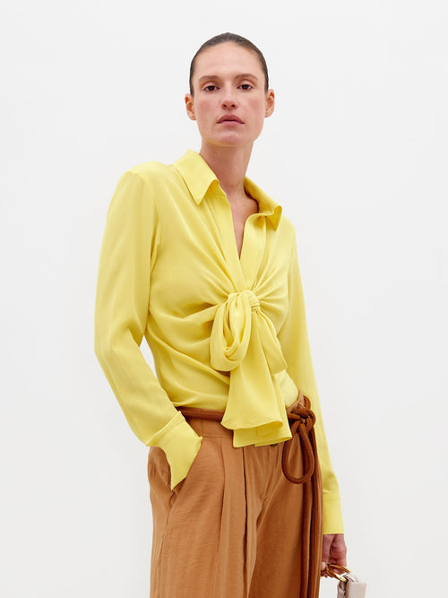 An individual stands against a plain backdrop wearing the Mercury Blouse Yellow, featuring long sleeves and a knotted front, paired with light brown belted trousers. They hold a small beige bag in their left hand. Pre-order now to ensure delivery by February 15th, 2025.