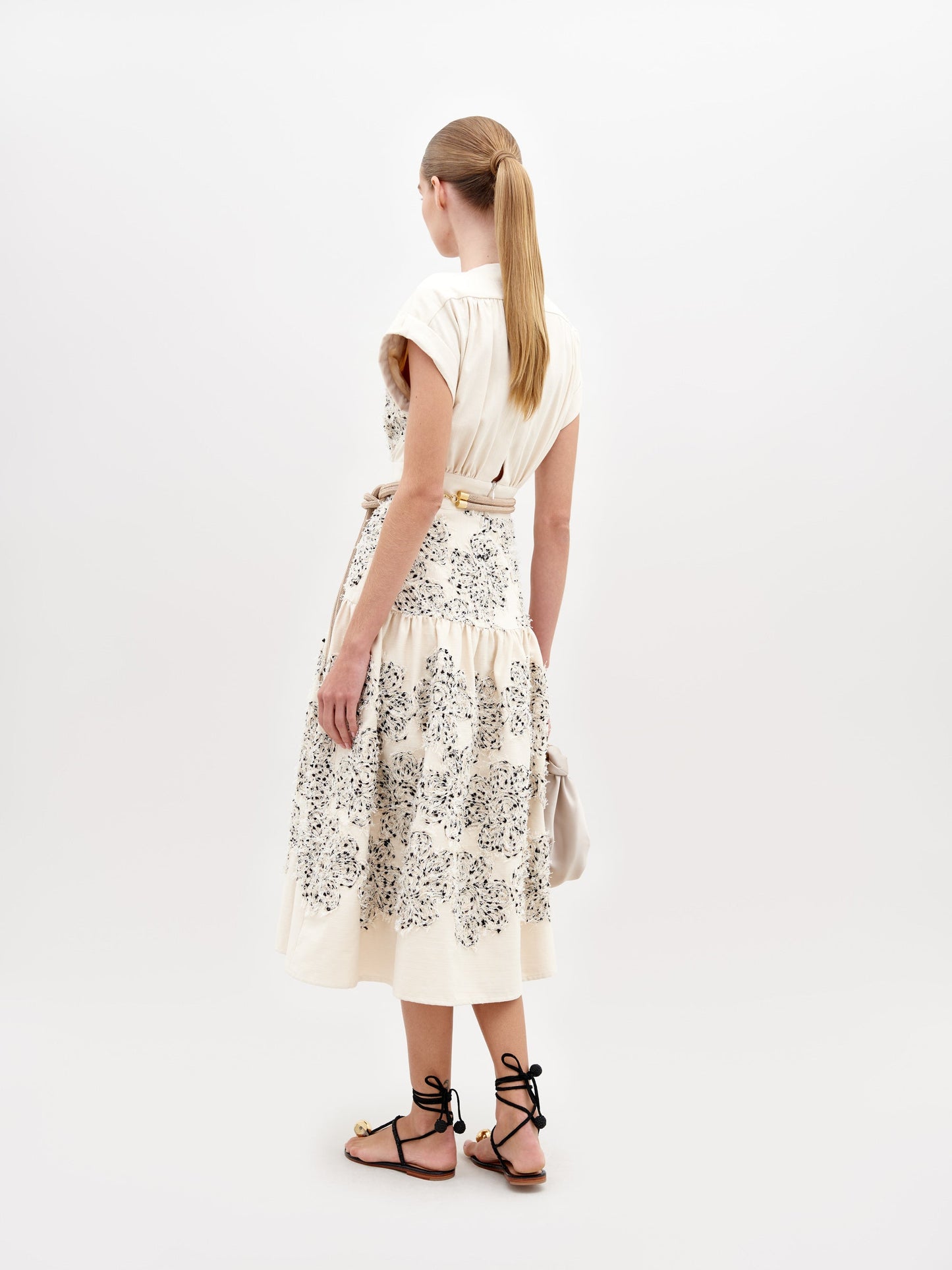 A woman wearing a Metaponto Dress Beige Black Embroidered Flowers with a beige belt and black sandals stands against a plain white background, ready for the fashion pre-order set to ship by date November 15th, 2024.