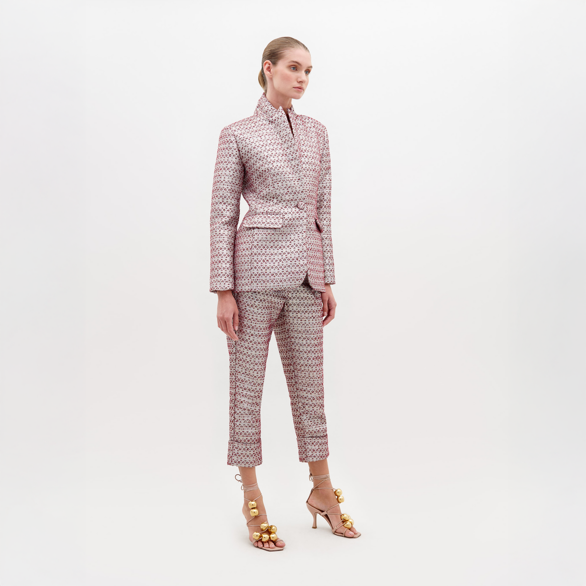 A woman stands against a plain white background wearing SVT DESIGNS’ Moad Pant in burgundy, a tailored Italian textile with a cropped, high-waisted fit, paired with gold-embellished high heels. Her hair is pulled back and she looks slightly to the side.